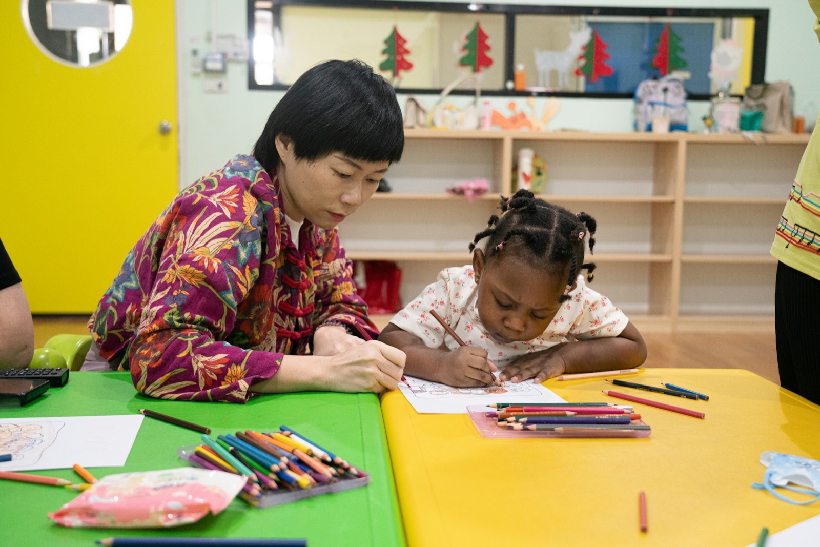 A teacher and a girl with pigtails coloring at a classroom table